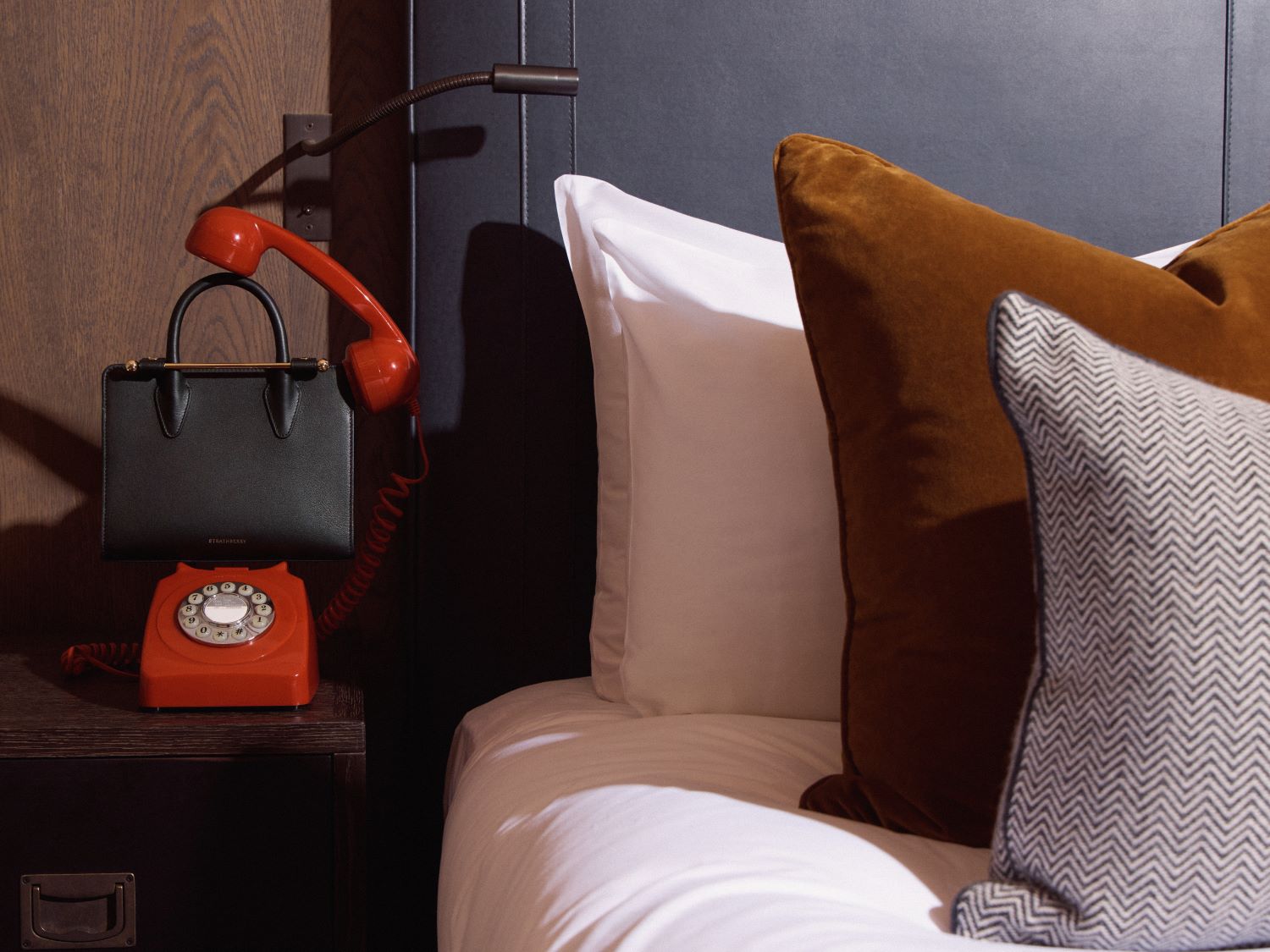 Hotel bed with bedside table featuring a Strathberry handbag in between a British red telephone