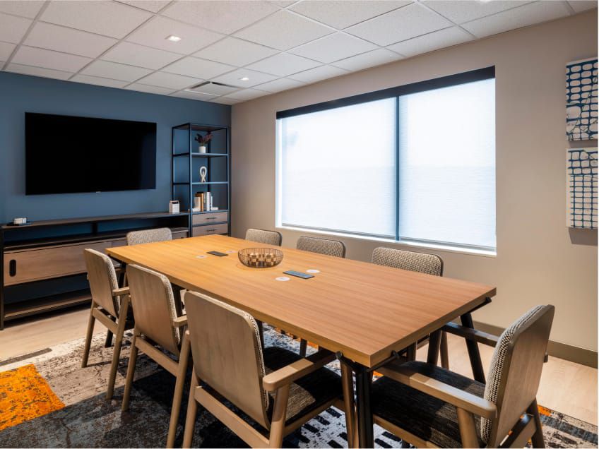 Warm and bright meeting room with a wood conference table, a striped rug, a flat screen monitor mounted to the wall and eight chairs.