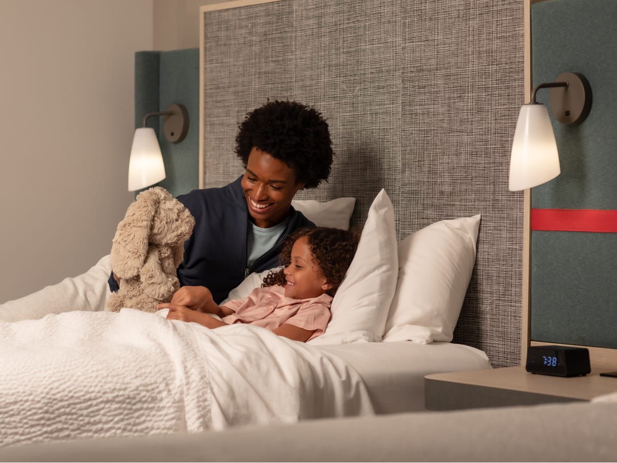 Mum tucking daughter into cosy hotel bed with stuffed animal