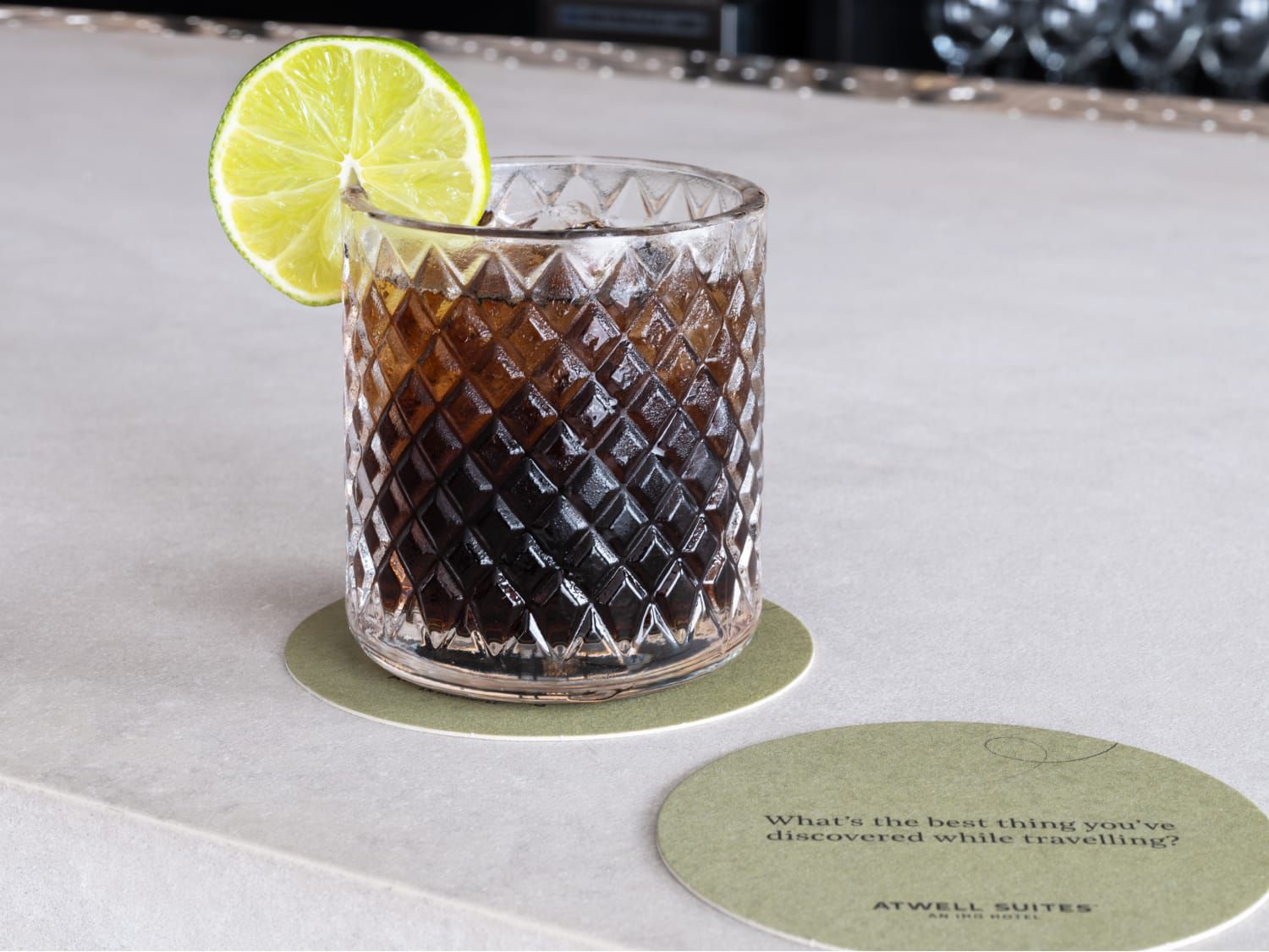 A textured glass filled with a dark drink, garnished with a lime slice, sits on a bar counter