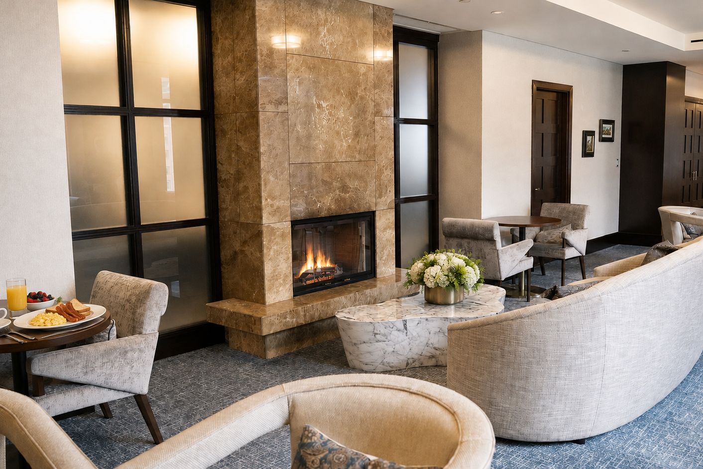 Elegant hotel club lounge with a marble fireplace, plush lounge seating, small dining tables, and a breakfast spread in a bright, contemporary setting.