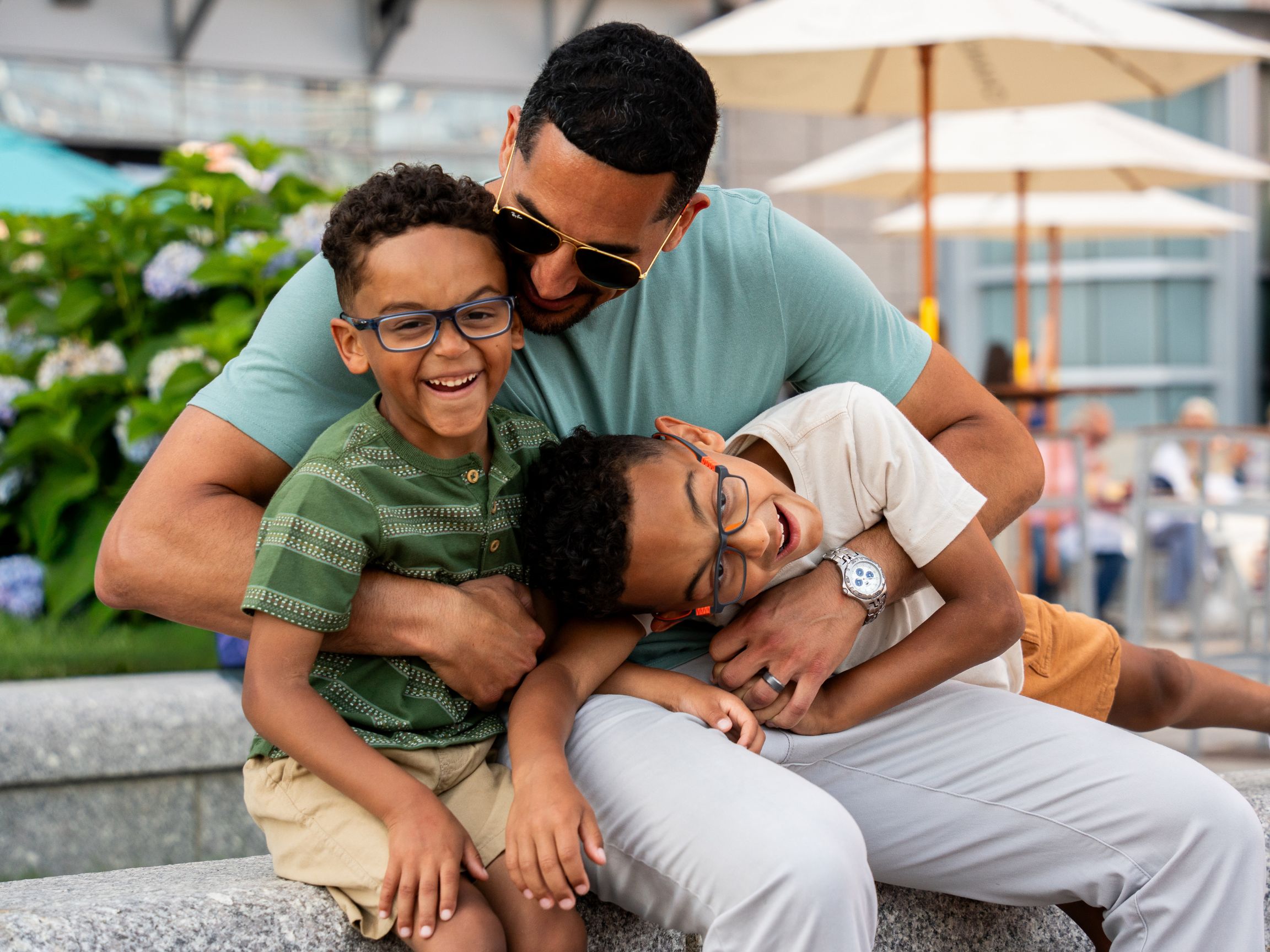 Adult seated outdoors with two children, sharing a playful embrace on a plaza bench with patio umbrellas and greenery in the background.