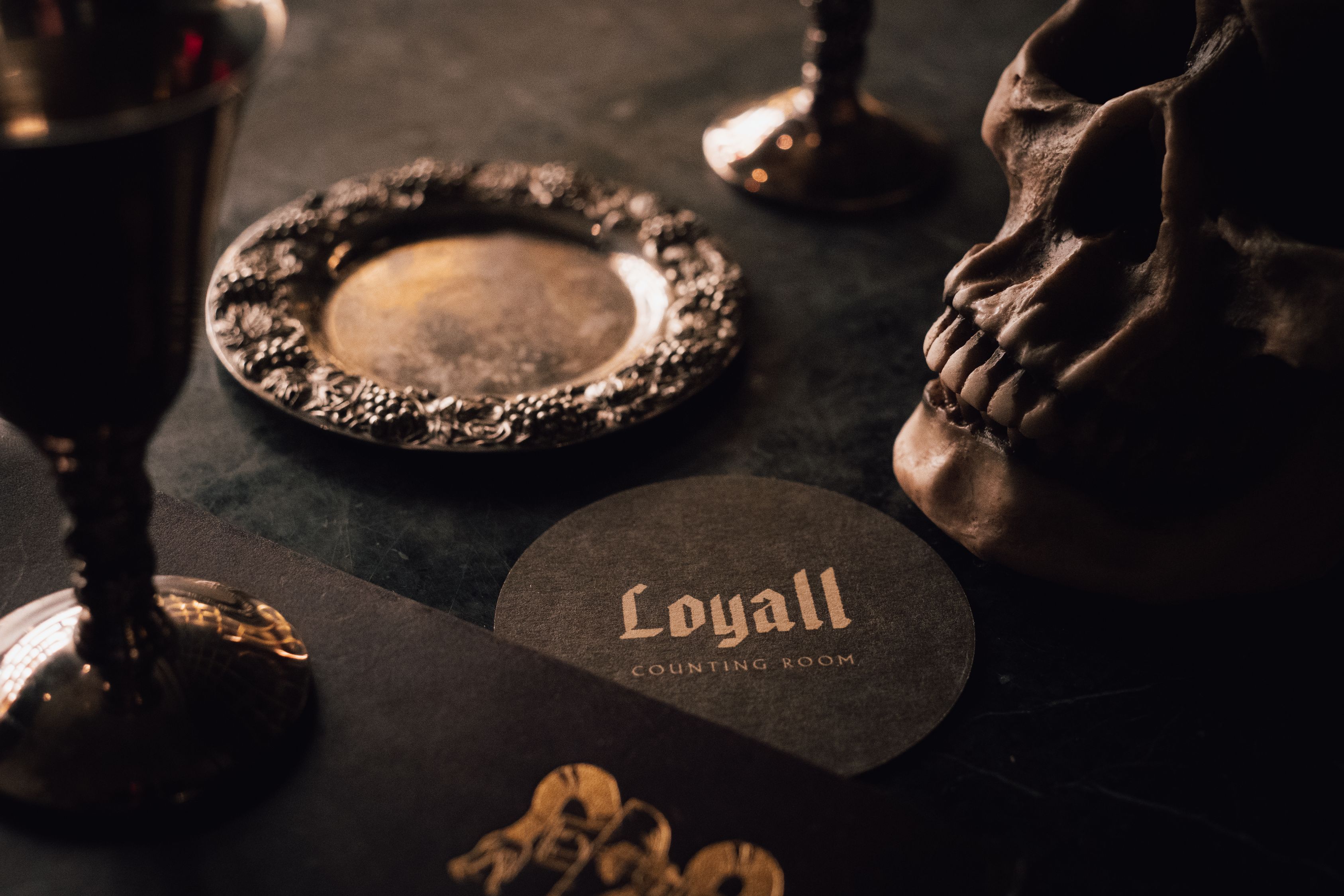 Dimly lit cocktail lounge display featuring decorative skulls, vintage-style glassware, and a Loyall Counting Room coaster on a dark tabletop.