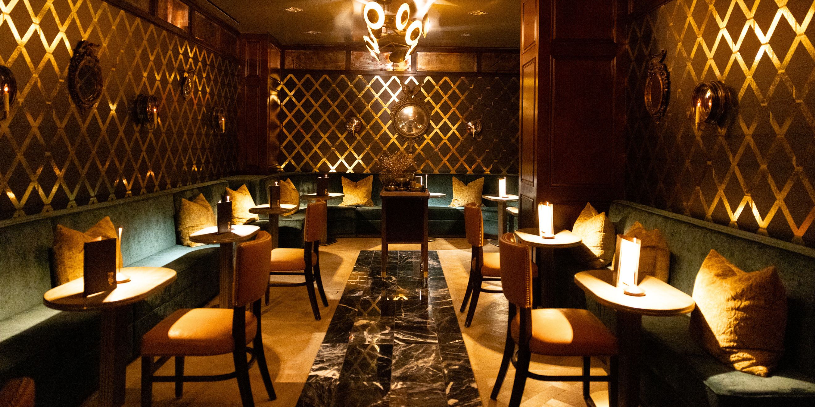 Intimate lounge with velvet banquette seating, candlelit tables, geometric gold wall accents, and warm ambient lighting creating an elegant cocktail bar atmosphere.
