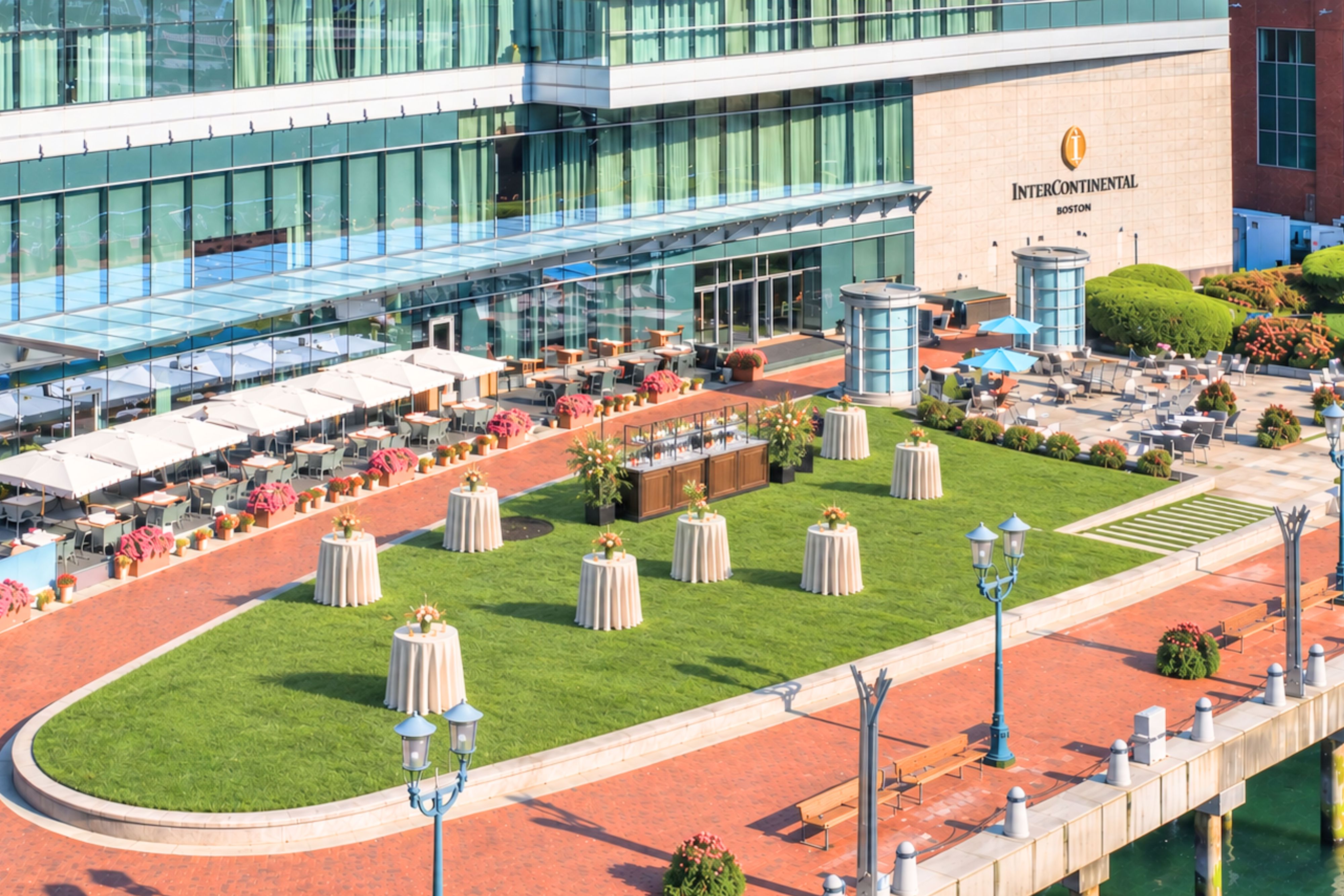 Outdoor event lawn at InterContinental Boston featuring cocktail tables, landscaped green space, patio seating, and a waterfront terrace beside the hotel's glass façade.