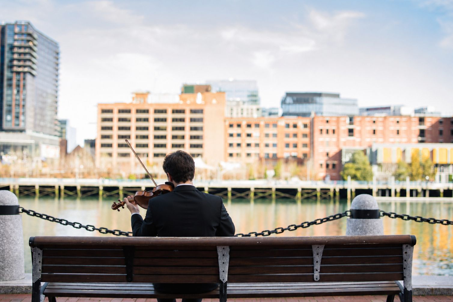 Man playing the violin on a bench in front of the harbor.