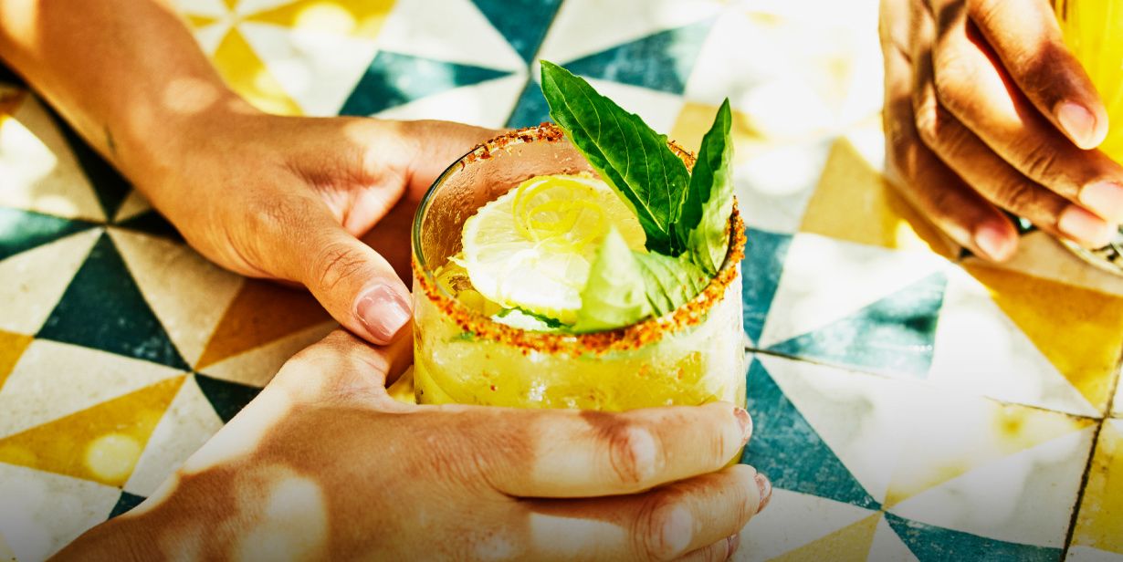 Two hands holding colourful cocktails over a brightly tiled table
