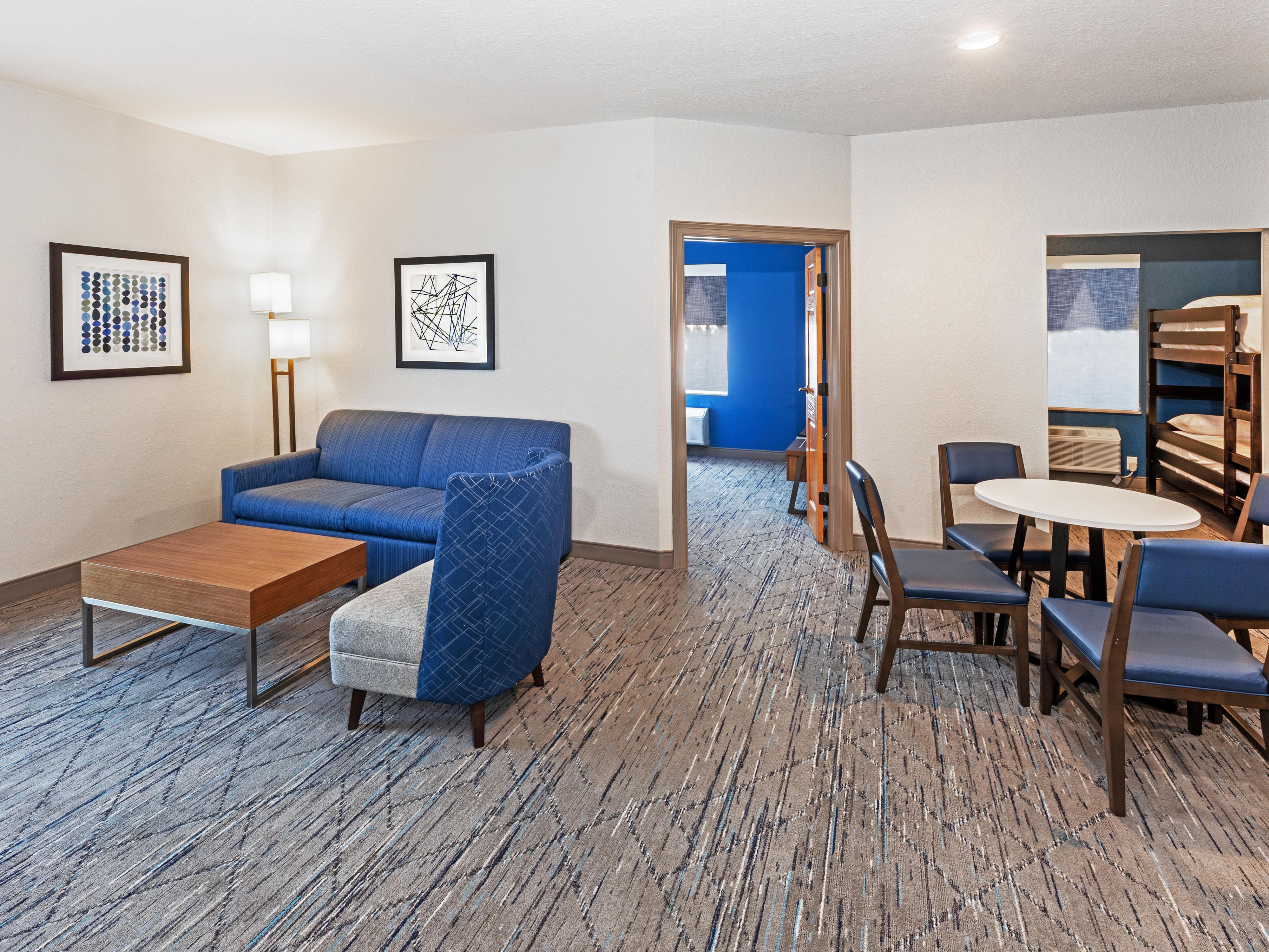 The Family Suite features an open floor plan with a dining & living area, & kitchenette equipped with a minifridge and microwave. The king bed is located in a separate room with closing doors with its own TV. A second area includes twin-size bunk beds that integrate with the open layout with tv. The living area features a sleeper sofa.