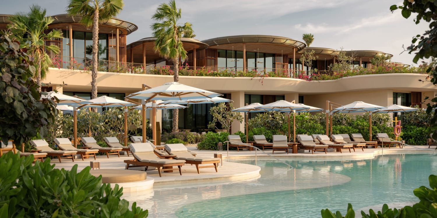 An architectural view of the InterContinental Red Sea Resort, featuring modern curved wooden roofs, flower-lined balconies, and striped beach umbrellas under palm trees.