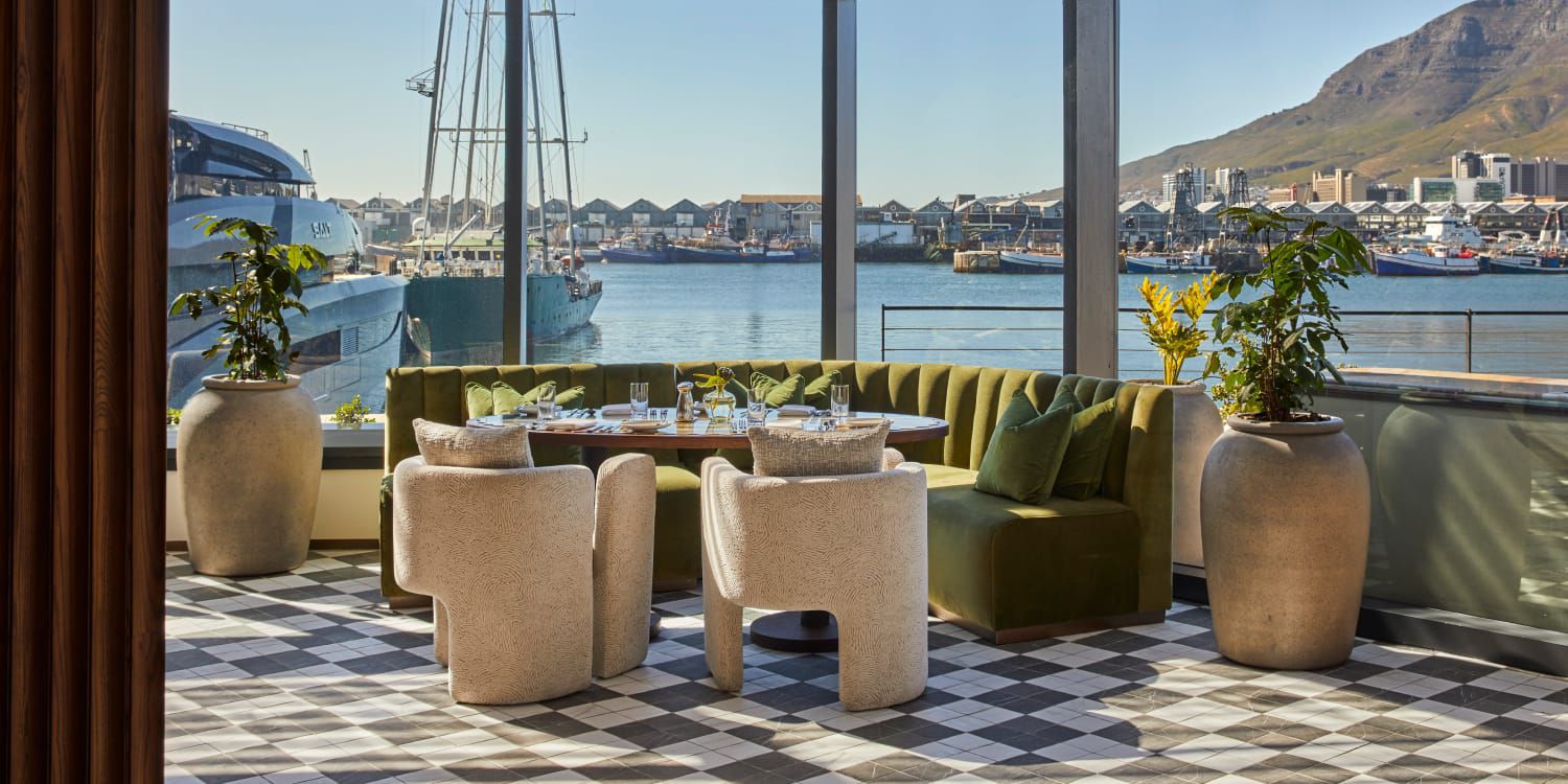 A sunlit modern restaurant terrace featuring a green velvet curved banquet, white textured armchairs, and checkered flooring, overlooking a vibrant harbor.