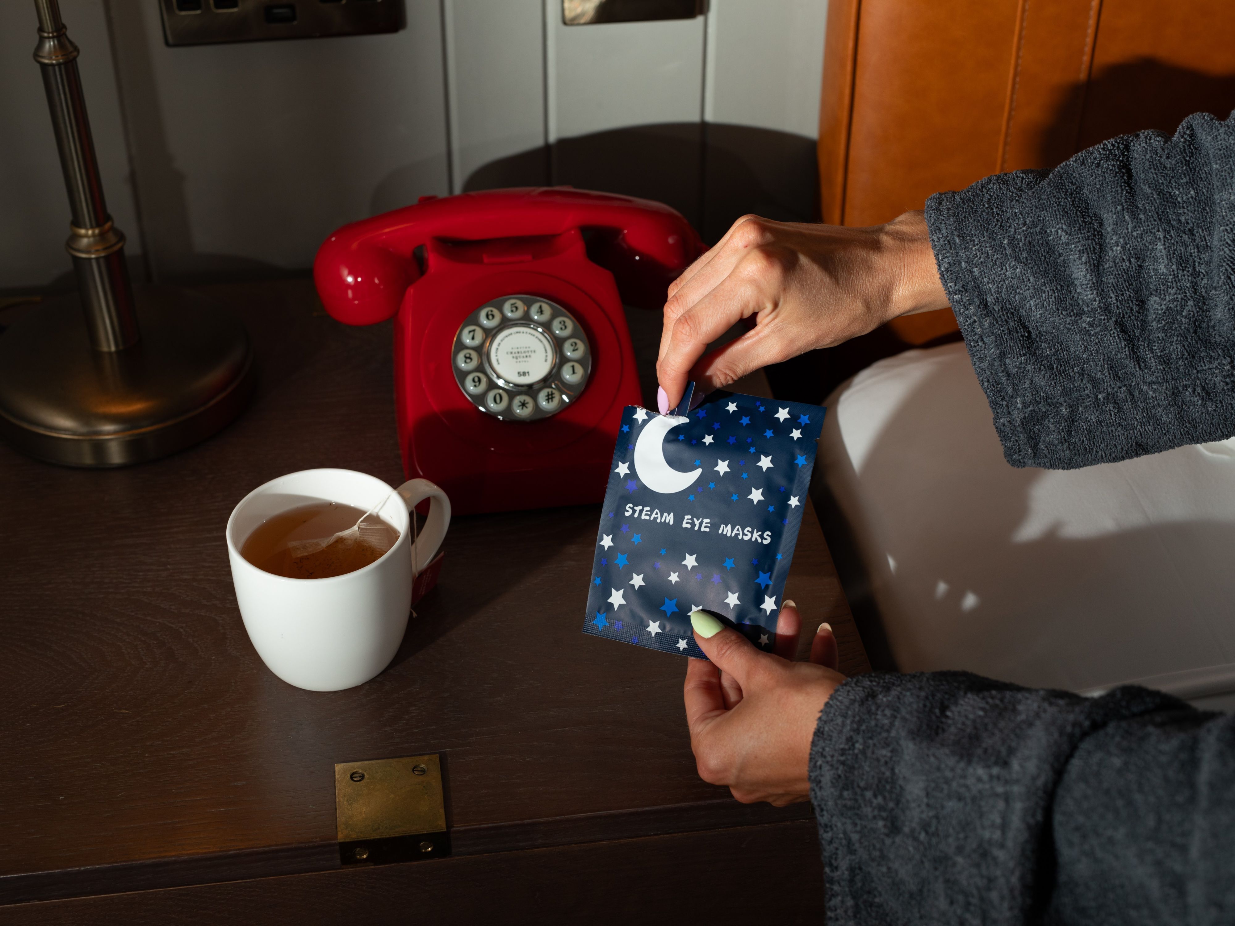 Hotel bedside table with added amenties featuring sleep mask, bath salts and a cupe of tea