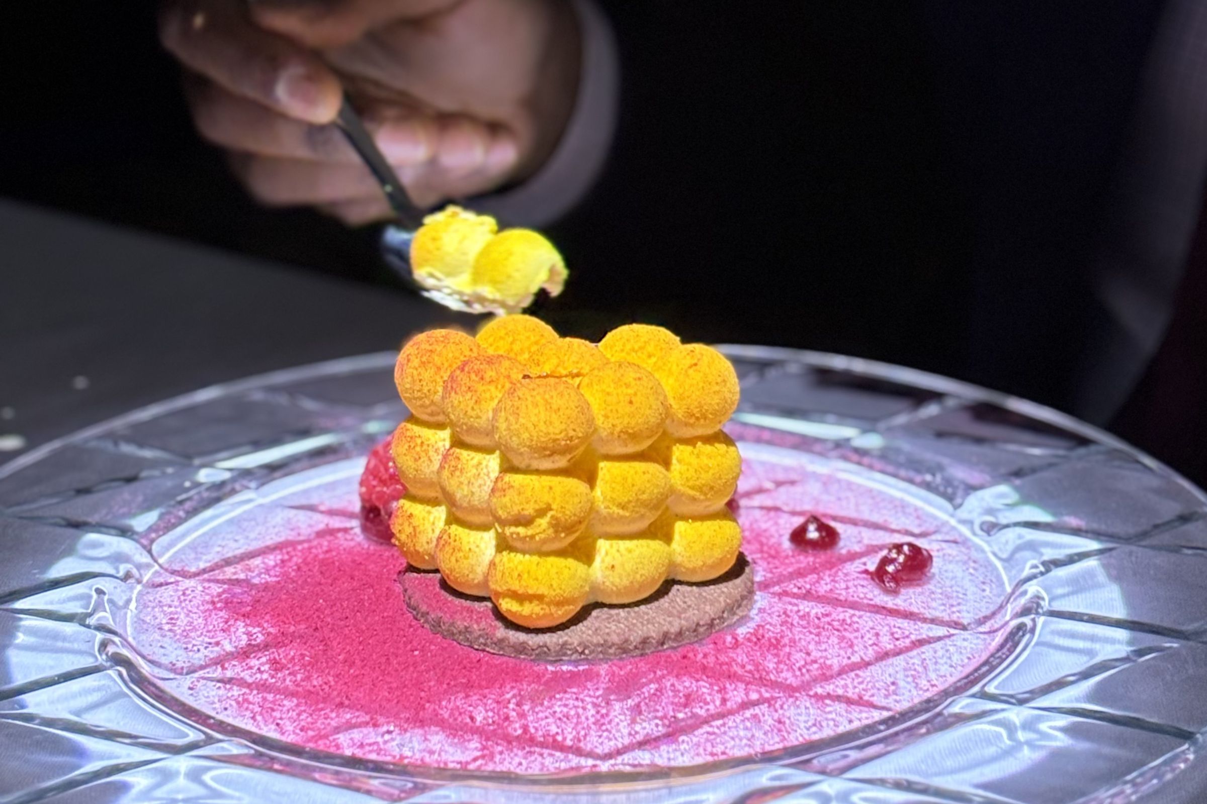 A plated gourmet dessert featuring a stack of yellow mousse spheres atop a cookie base with pink powdered garnish.
