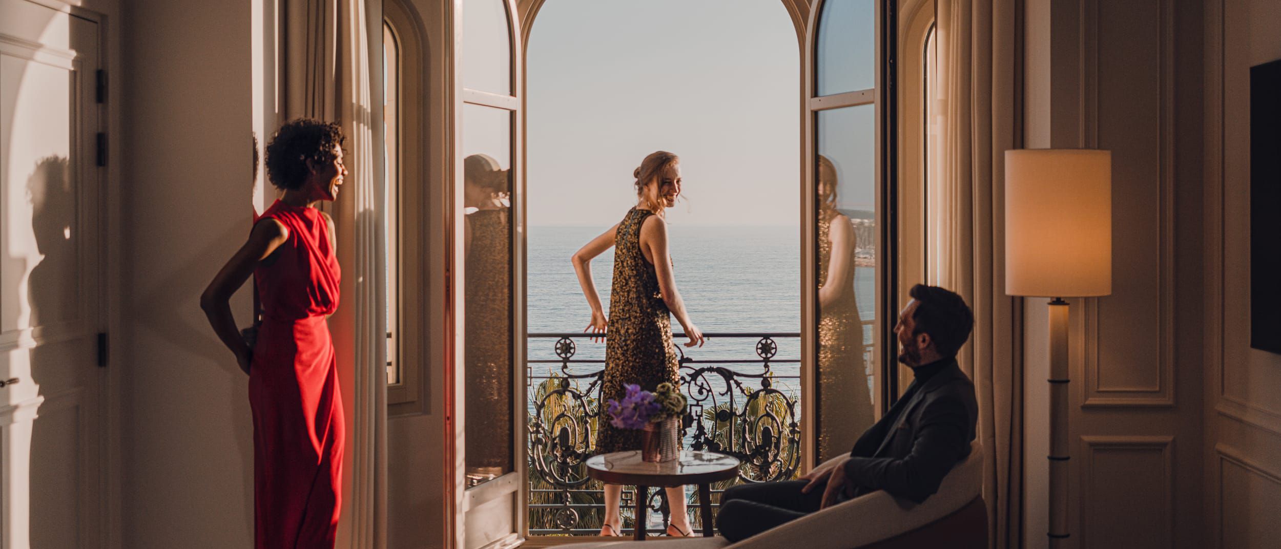 Two well-dressed women and a gentleman looking out over a crystal sea from the balcony of a suite.