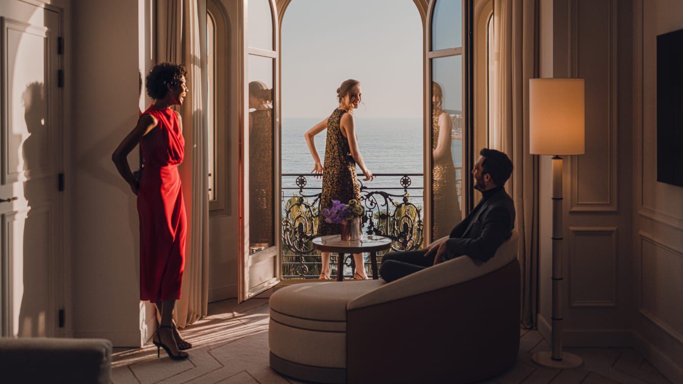 Two well-dressed women and a gentleman looking out over a crystal sea from the balcony of a suite.