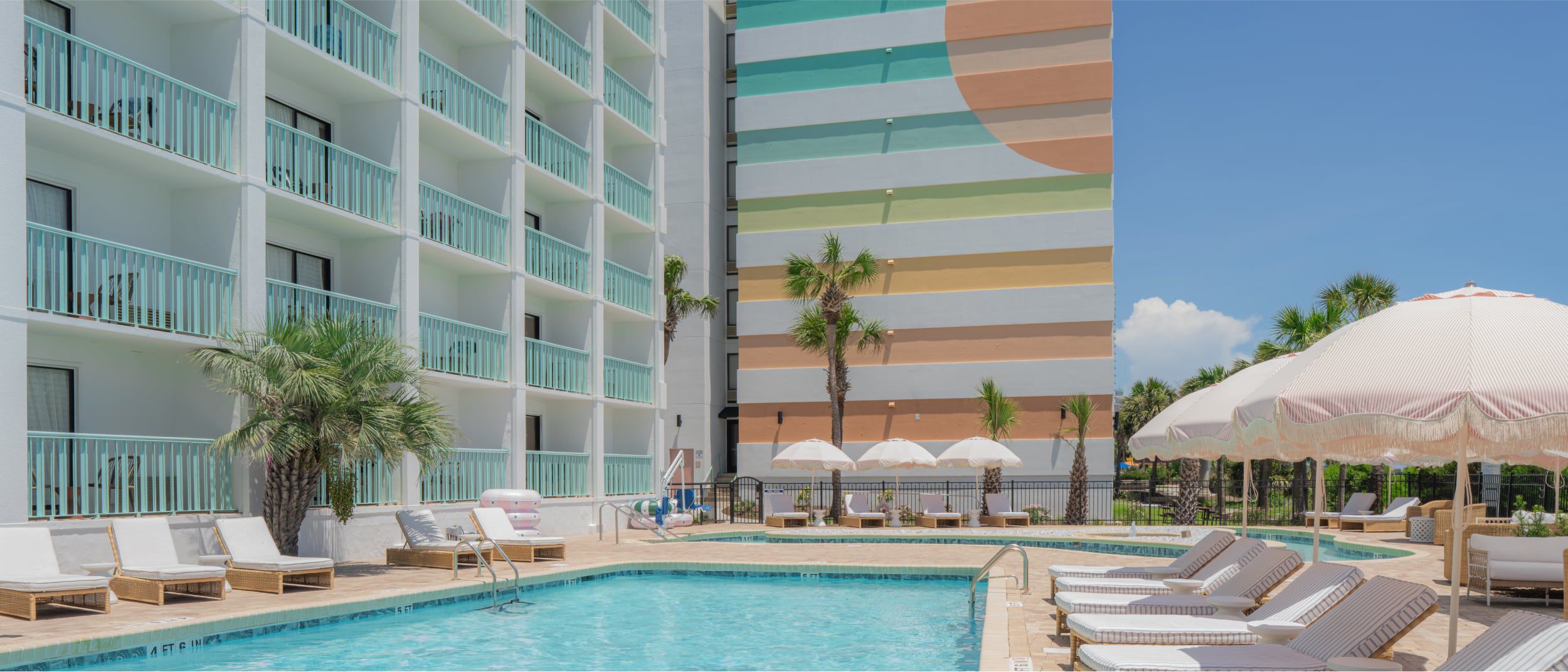 An outdoor pool scene with wicker loungers and umbrellas against a pastel building backdrop.